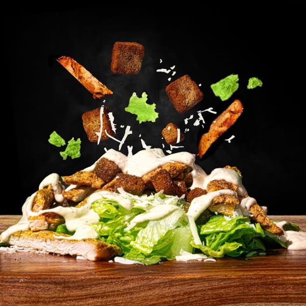 Caesar salad with grilled chicken, croutons, and dressing falling onto a wooden surface.