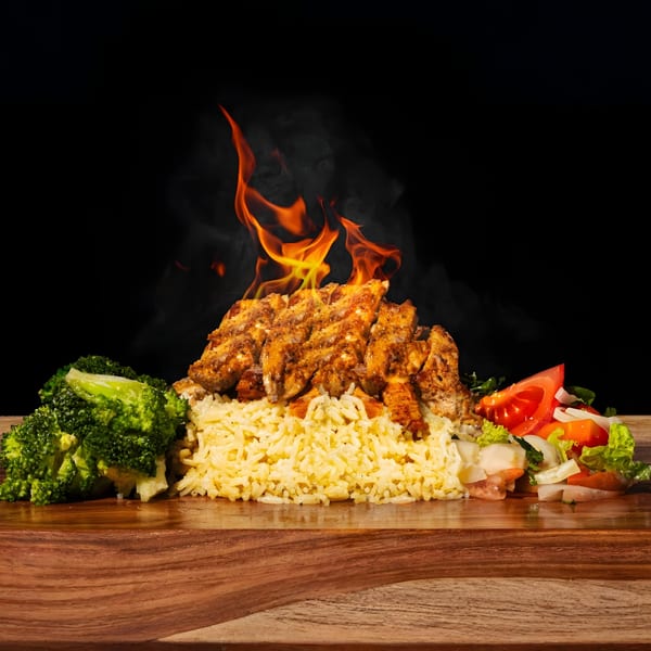Flaming grilled chicken over rice, with broccoli and a side salad on a wooden board.