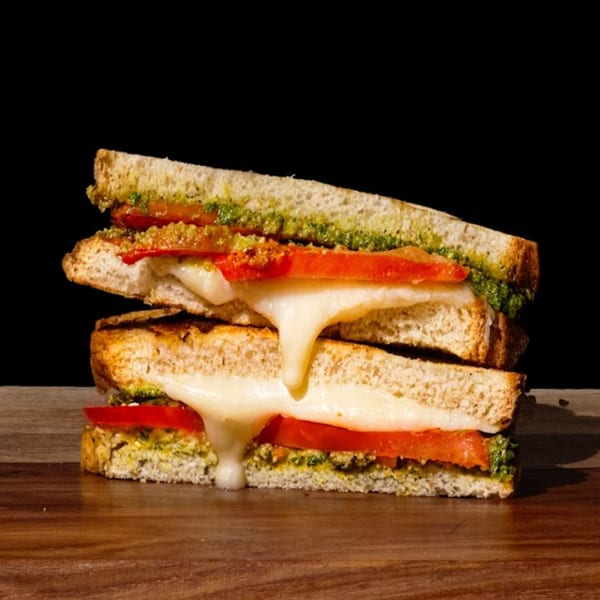 A grilled cheese sandwich with pesto and tomato, cheese oozing out, on a dark wooden surface.